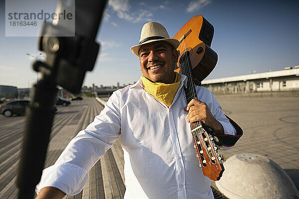 Lächelnder Gitarrist hält Gitarre und macht an einem sonnigen Tag ein Selfie mit dem Smartphone