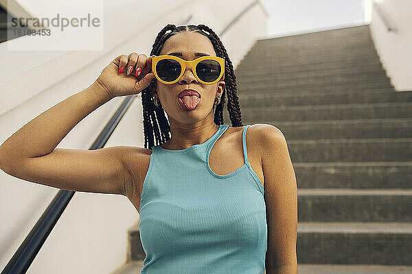 Junge Frau mit Sonnenbrille streckt auf der Treppe die Zunge heraus