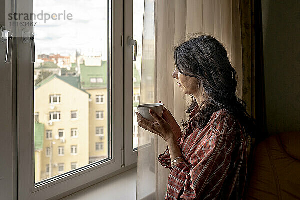 Frau hält Kaffeetasse in der Hand und schaut zu Hause durch das Fenster