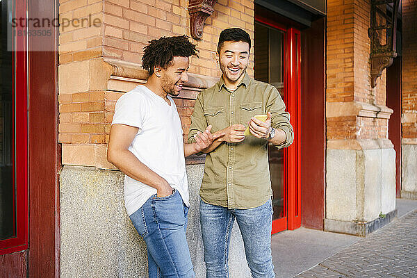 Young multiracial men laughing and using phone together on footpath