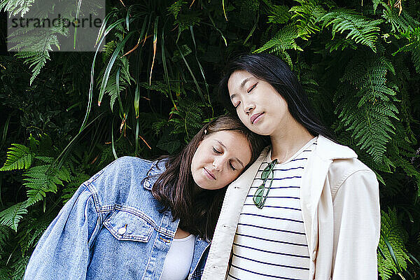 Lesbian women with eyes closed leaning on each other in front of plants