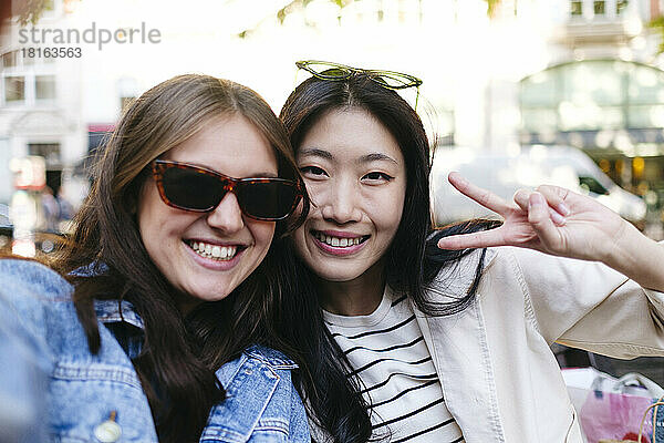 Happy woman wearing sunglasses taking selfie with lesbian friend showing peace sign