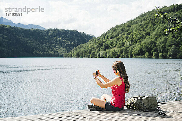 Frau sitzt am Steg und fotografiert den Levico-See per Smartphone