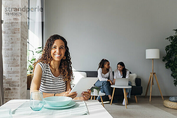 Smiling woman holding tablet PC with friends in background at home