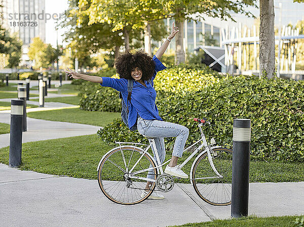 Glückliche Afro-Frau mit erhobenen Armen sitzt auf dem Fahrrad im Park