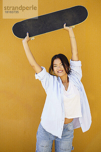 Glückliche junge Frau hält Skateboard vor gelber Wand hoch