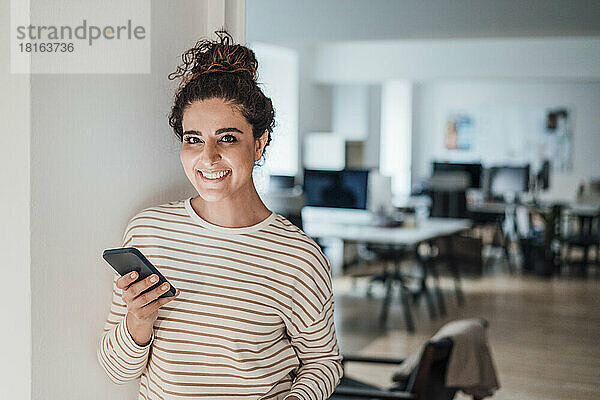 Lächelnde Geschäftsfrau mit Smartphone an der Wand im Büro