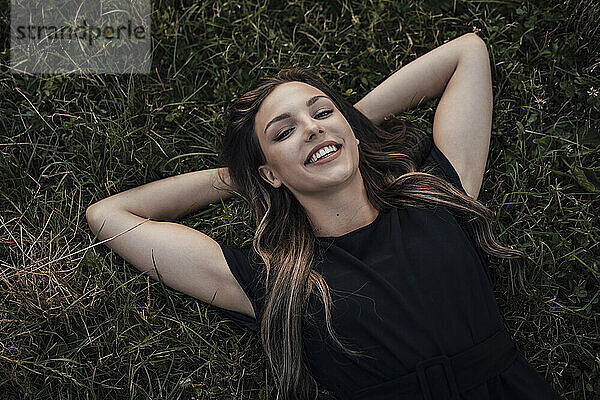 Glückliche junge Frau mit den Händen hinter dem Kopf entspannt sich im Gras im Park