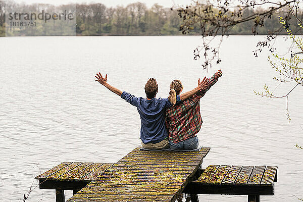 Carefree couple with arms outstretched sitting on jetty at lake
