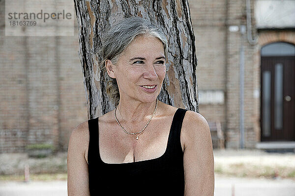 Smiling beautiful senior woman leaning on tree trunk