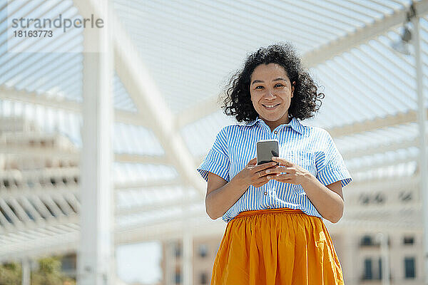 Lächelnde Frau mit Mobiltelefon steht an einem sonnigen Tag