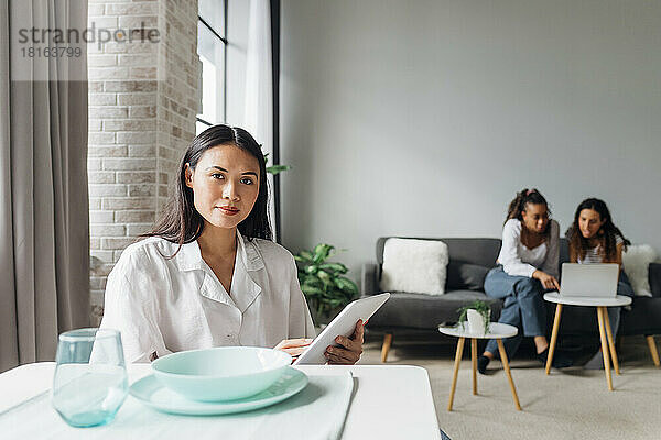Confident woman holding tablet PC with flatmates in background at home