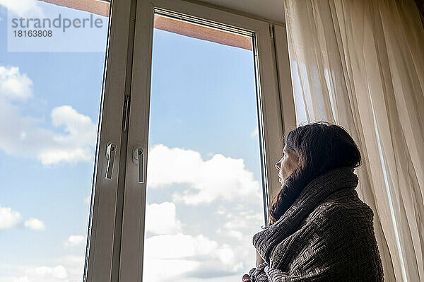 Nachdenkliche Frau  in eine Decke gehüllt  blickt zu Hause durch das Fenster