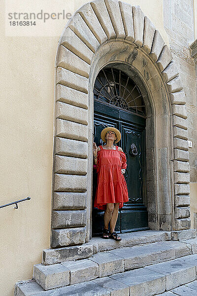 Ältere Frau in rotem Kleid und Sonnenhut steht vor alter Tür