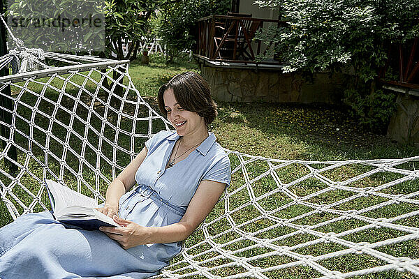 Smiling woman reading book sitting on hammock at sunny day