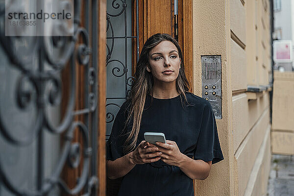 Nachdenkliche junge Frau mit Smartphone steht vor der Tür
