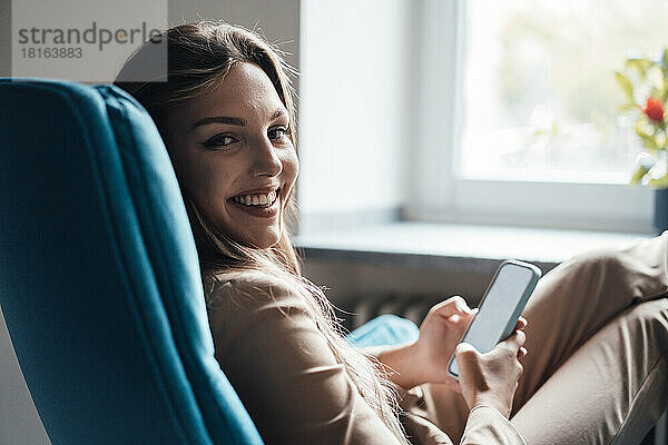 Smiling young businesswoman with smart phone sitting on chair at home