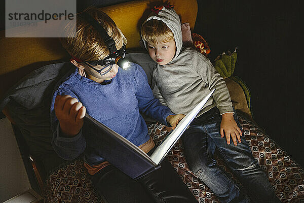 Boy wearing headlight reading book with brother on bed at home