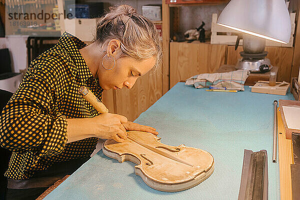 Gitarrenbauer mit Handwerkzeugschnitzerei an Geige am Schreibtisch in der Werkstatt