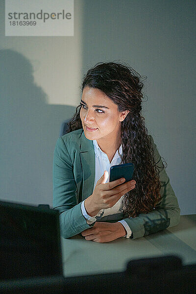 Nachdenkliche Geschäftsfrau mit Mobiltelefon am Schreibtisch im Büro