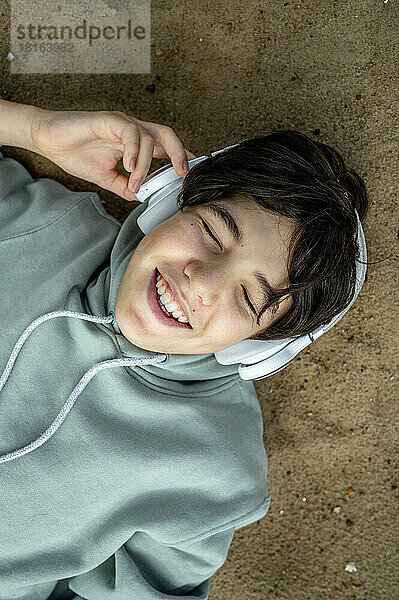 Fröhlicher Junge mit kabellosen Kopfhörern  der Musik hört und sich am Strand entspannt