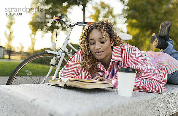 Junge Frau mit der Hand im Haar liest Buch auf Bank