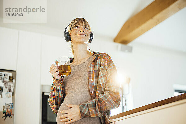 Lächelnde schwangere Frau mit Kopfhörern  die zu Hause eine Teetasse hält