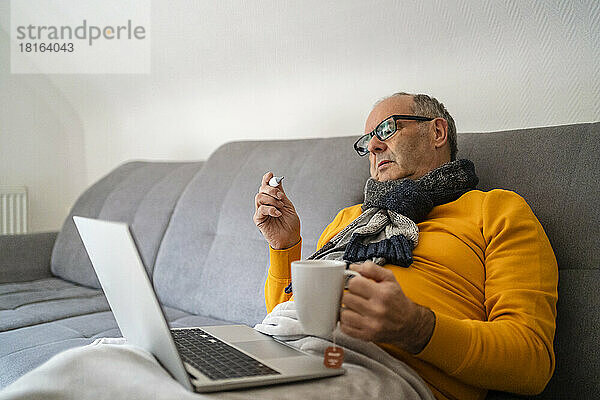 Mann mit Laptop hält Becher und prüft Fieber durch Thermometer auf Sofa