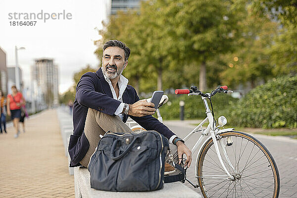 Glücklicher Geschäftsmann mit Smartphone  der sich mit dem Fahrrad auf einer Bank ausruht