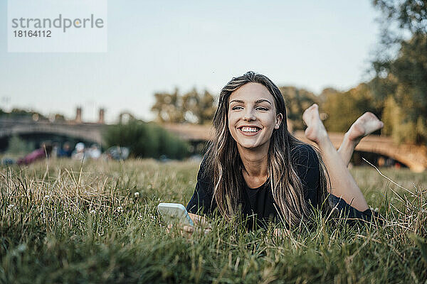 Glückliche Frau mit Smartphone liegt im Gras im Park