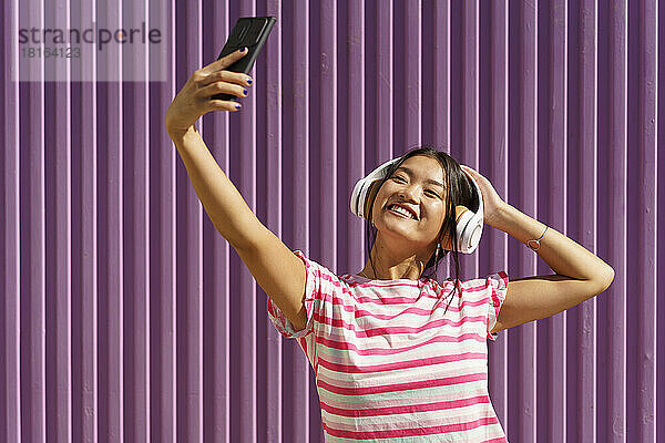 Lächelnde junge Frau macht ein Selfie mit ihrem Smartphone vor einer Wellblechwand