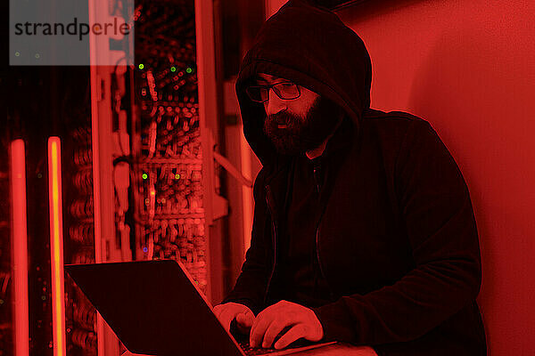 Hacker trägt Kapuzenpullover und programmiert auf Laptop im Serverraum