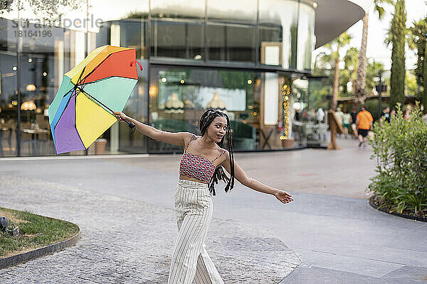 Junge Frau mit ausgestreckten Armen hält bunten Regenschirm auf dem Fußweg