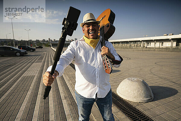 Fröhlicher Straßenmusiker hält Gitarre und macht ein Selfie mit dem Mobiltelefon auf einem Stativ