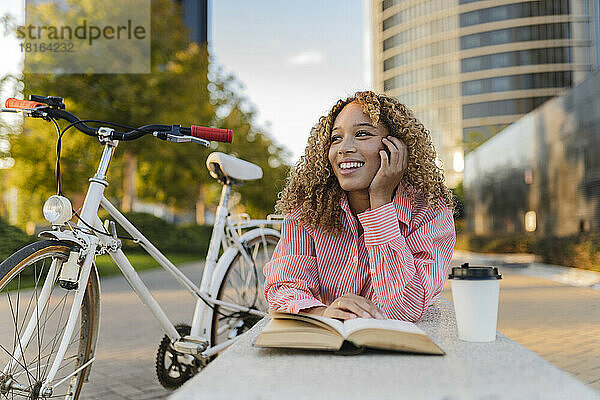 Lächelnde Frau mit Buch und Einwegkaffeetasse auf Bank liegend