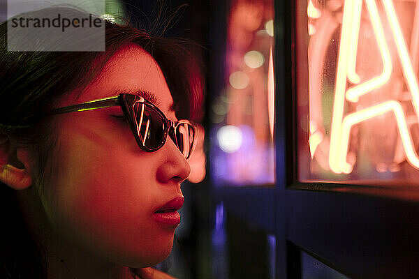 Young woman wearing sunglasses by illuminated wall with neon lights