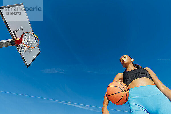 Nachdenklicher Basketballspieler mit Ball am Reifen an einem sonnigen Tag