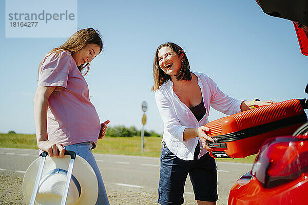 Glückliche Frau lädt Koffer in den Kofferraum eines Autos und schaut schwangere Freundin an