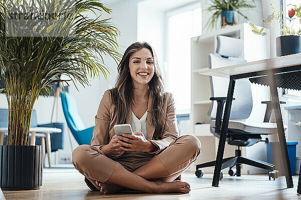 Glückliche junge Geschäftsfrau mit Mobiltelefon sitzt im Heimbüro