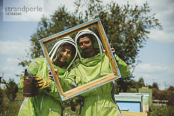Smiling beekeepers looking through wooden frame at apiary