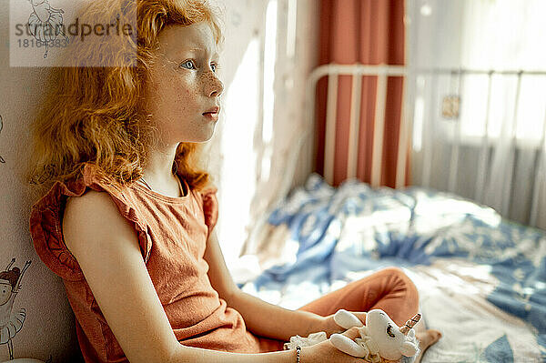Nachdenkliches rothaariges Mädchen mit Stofftier sitzt auf dem Bett
