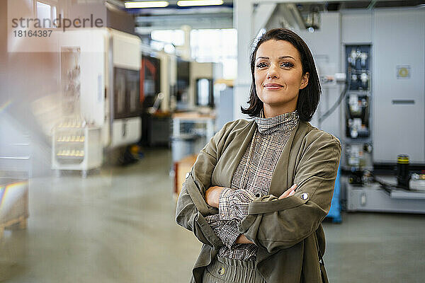 Lächelnde Geschäftsfrau mit verschränkten Armen steht in der Fabrik