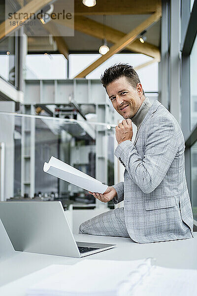Lächelnder Geschäftsmann hält Papier und sitzt mit der Hand am Kinn am Laptop am Schreibtisch