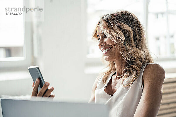 Lächelnde Geschäftsfrau blickt im Büro auf ihr Smartphone