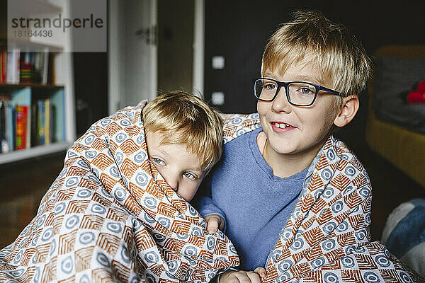 Happy boy with brother wrapped in blanket sitting at home