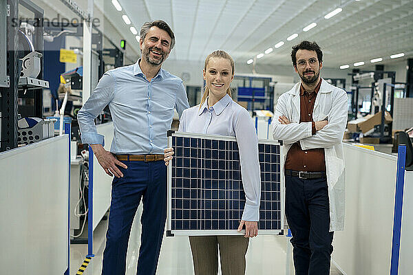 Lächelnder junger Ingenieur hält Solarpanel in der Hand und steht mit Kollegen in der Fabrik