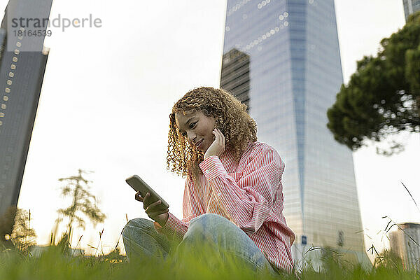 Lächelnde junge Frau benutzt Mobiltelefon auf Gras in der Stadt