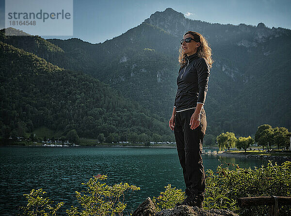 Reife Frau mit Sonnenbrille steht auf einem Felsen am See