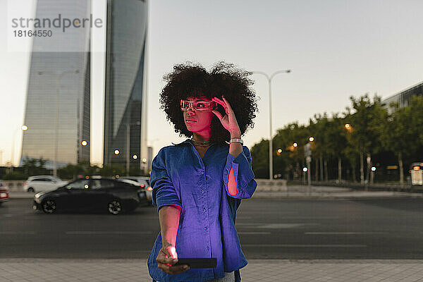 Rotes Licht fällt auf das Gesicht einer Afro-Frau  die eine intelligente Brille trägt und ihr Smartphone auf der Straße hält