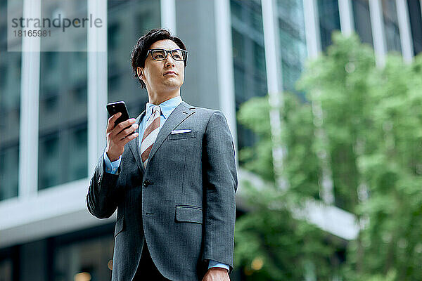 Japanischer Geschäftsmann hält ein Smartphone in der Hand und schaut weg